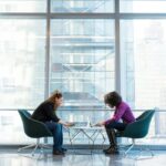 two women against a glass wall in an office environment working on laptop computers