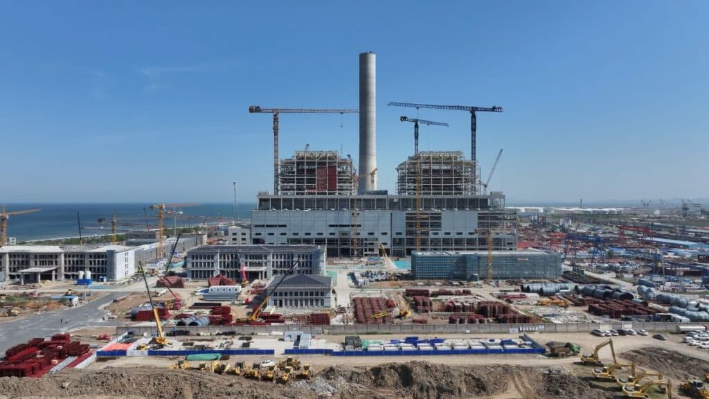 Large-scale thermal power plant under construction with cranes, steel framework, and coastal background